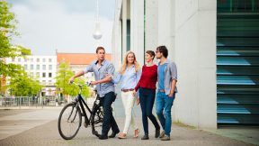 Vier Studierende gehen durch Berlin mit einem Fahrrad, im Hintergrund sieht man den Fernsehturm.